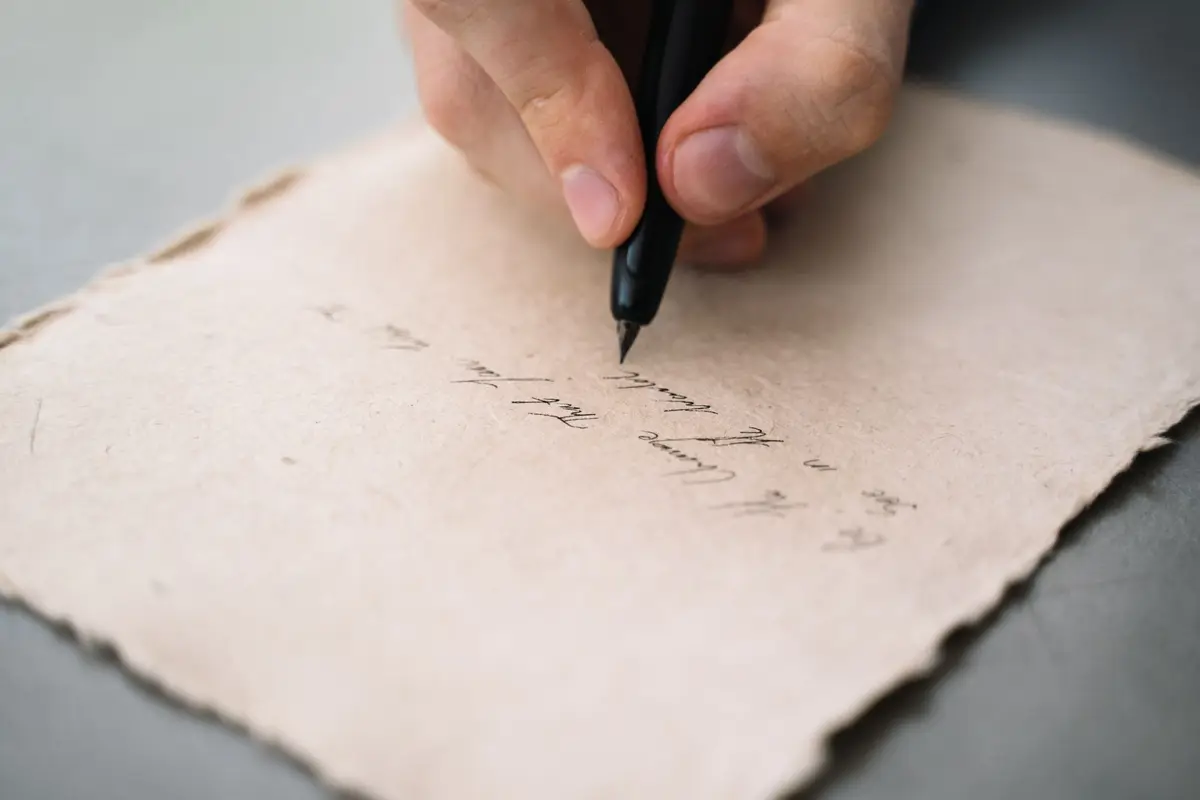 A hand writing a letter in ink on deckle-edged paper