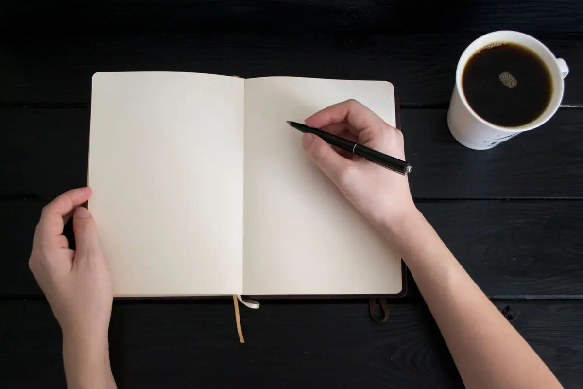 Hands writing in a blank notebook beside a cup of coffee
