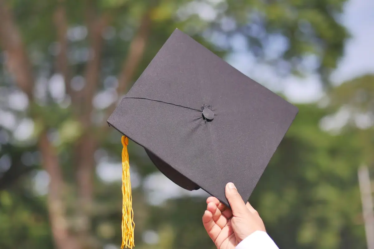 A hand holding up a graduation cap against green trees