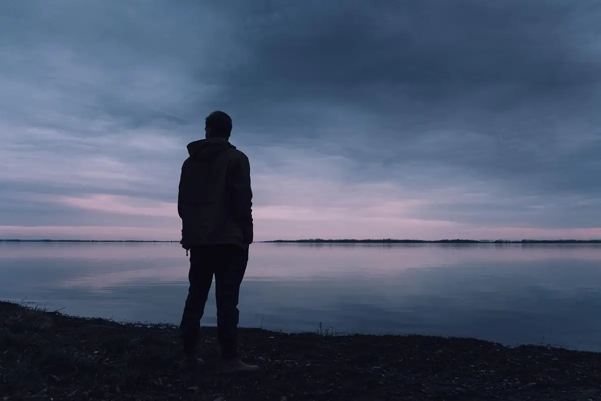 A person standing alone at the edge of a still lake at dusk